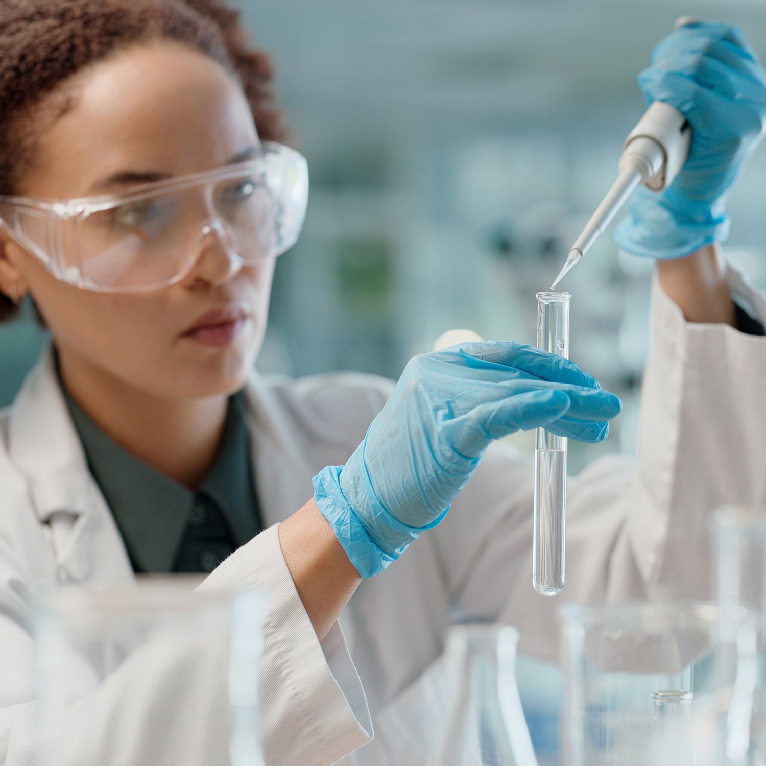 Scientist in a lab wearing goggles and gloves, working with a test tube and pipette.